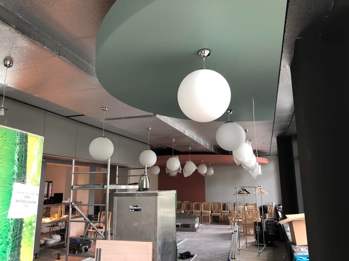 Wnętrze lokalu gastronomicznego w trakcie wykańczania, widoczne wiszące, kuliste lampy, częściowo jeszcze w folii ochronnej, surowy sufit z widoczną instalacją, krzesła ustawione w rzędach...