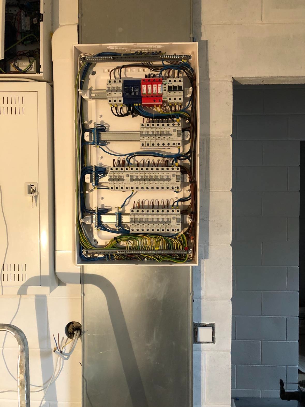 Otwarta skrzynka rozdzielcza z widocznymi bezpiecznikami, przewodami i listwami zaciskowymi, zamontowana na ścianie z surowych, pomalowanych bloków betonowych, obok metalowej rury i drugiej...