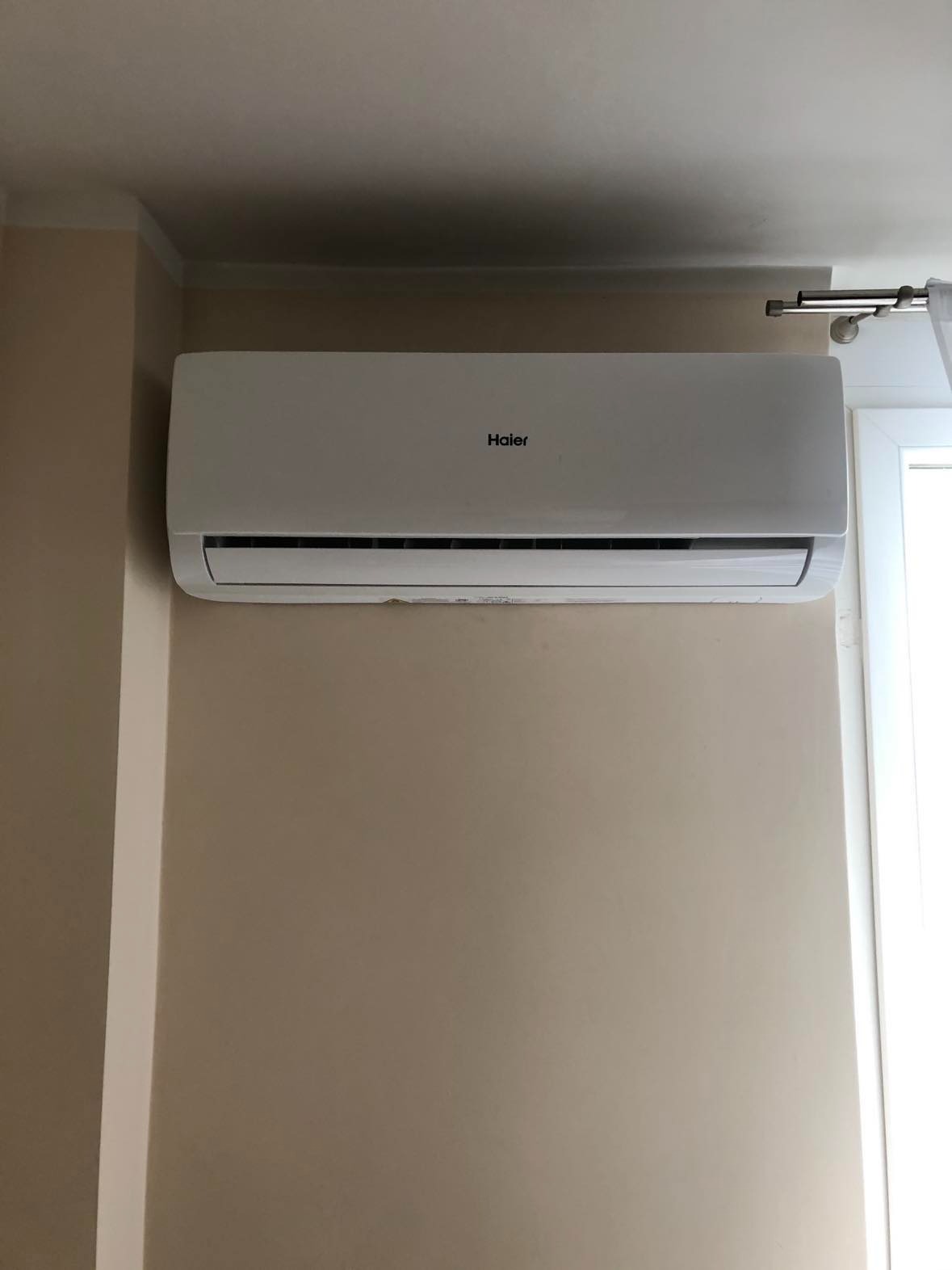 Klimatyzator Haier zamontowany na beżowej ścianie, widoczny fragment okna z zasłoną.