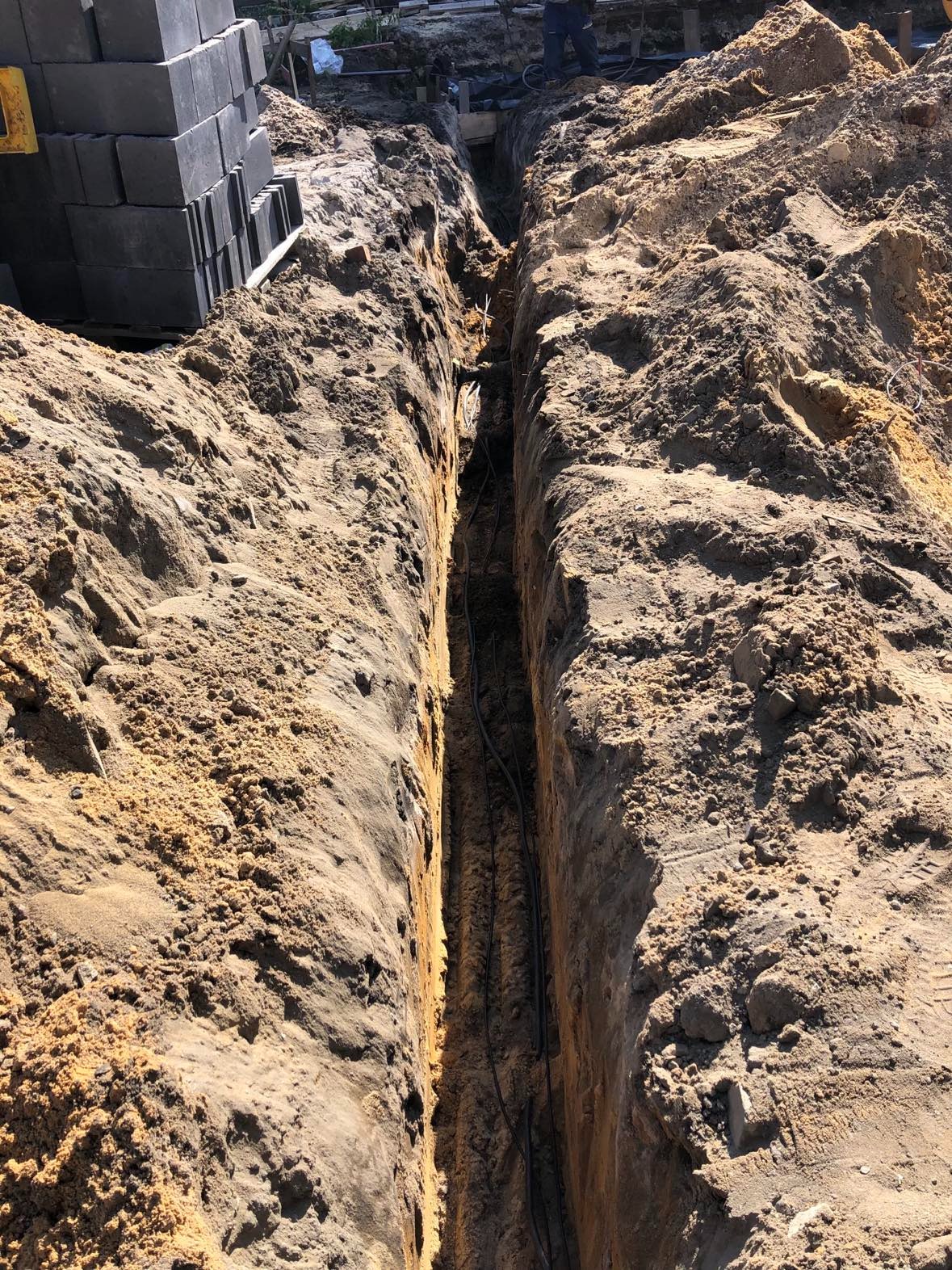 Wykop w ziemi z ułożonymi kablami elektrycznymi; obok stos szarych bloczków betonowych.