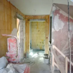 Wnętrze w trakcie prac wykończeniowych, widoczna maszyna do tynkowania, ściany częściowo pokryte tynkiem, widoczne przewody i narzędzia, surowy stan pomieszczenia.