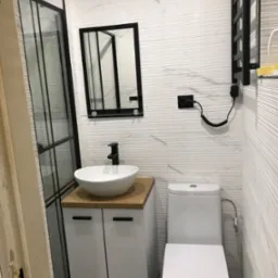 Nowoczesna łazienka z białą umywalką nablatową na szafce z drewnianym blatem, toaletą, grzejnikiem drabinkowym i kabiną prysznicową z czarnymi ramami, wykończona płytkami imitującymi marmur.