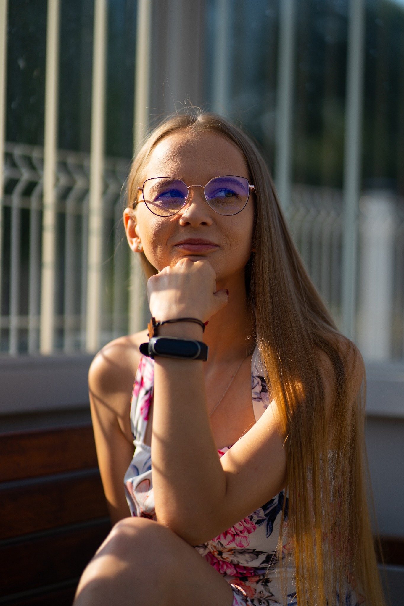 Portret młodej kobiety w okularach, opierającej podbródek na dłoni, siedzącej na ławce przed budynkiem z żaluzjami w tle.