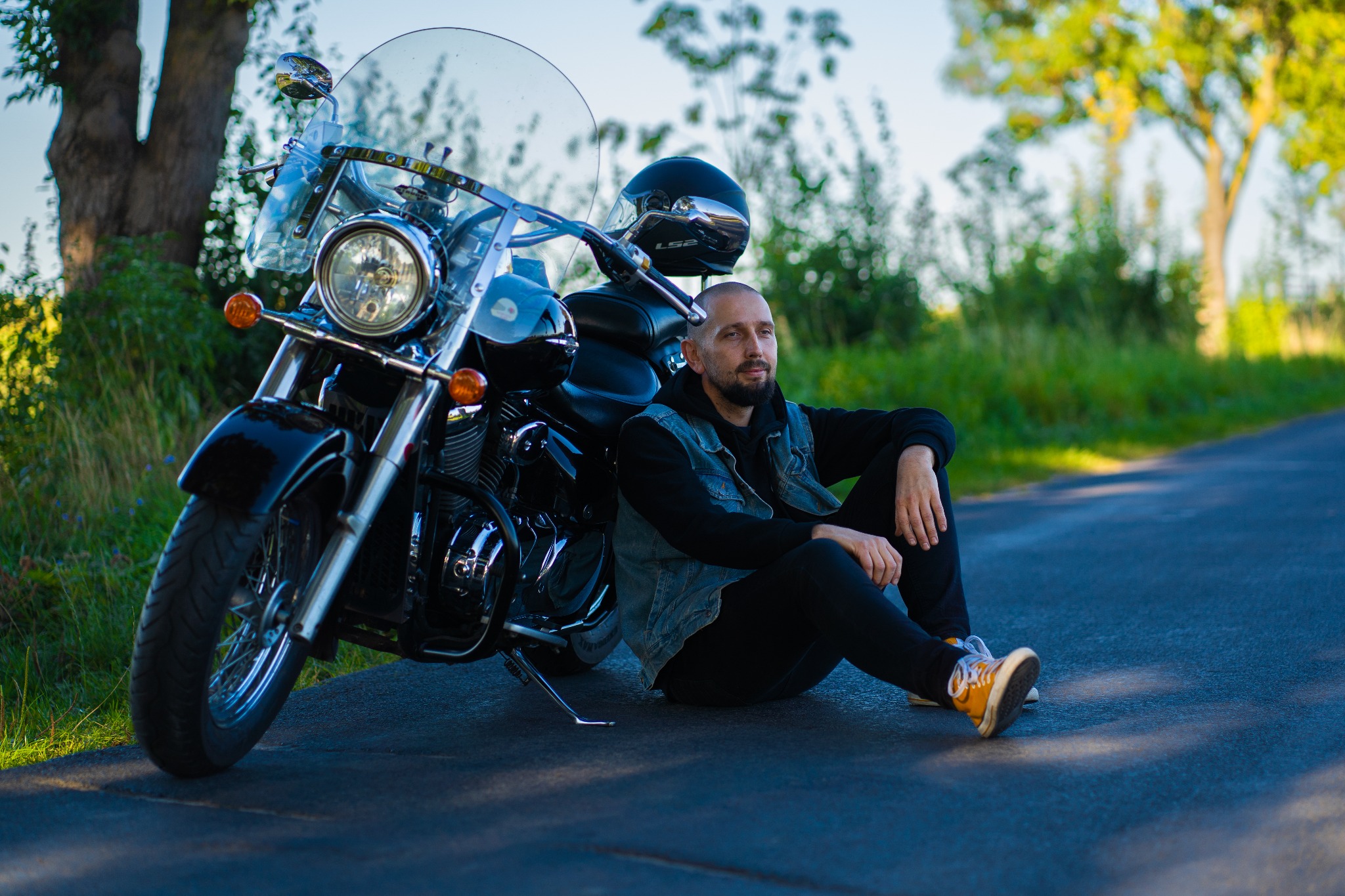 Mężczyzna w jeansowej kamizelce siedzi na asfalcie oparty o czarny motocykl z chromowanymi elementami, w tle drzewa i trawa.