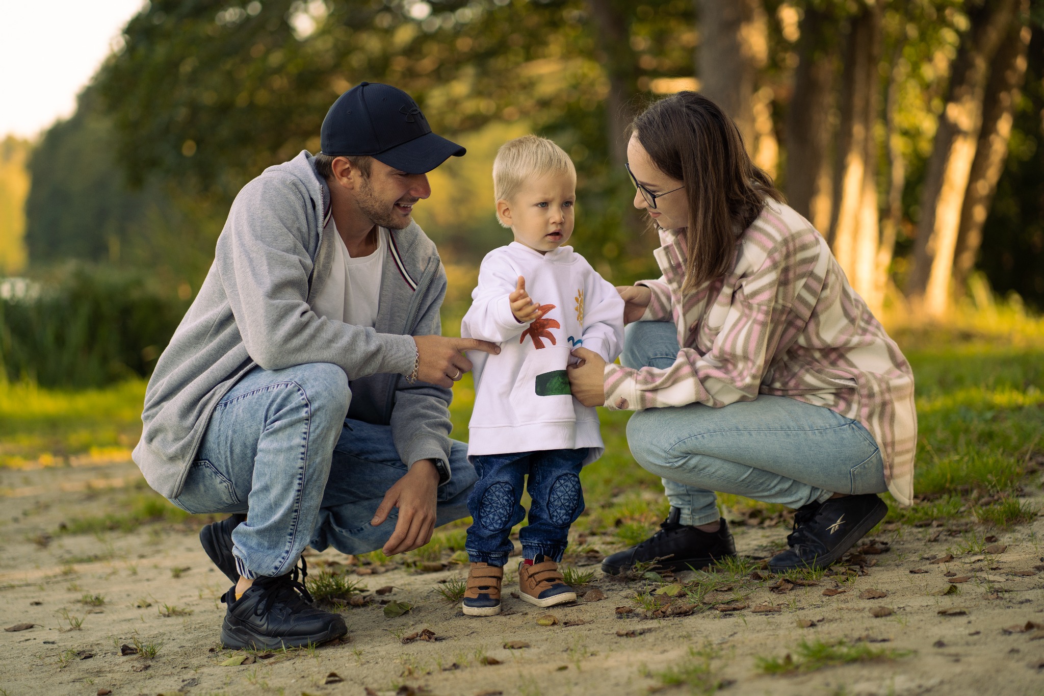 Rodzinne zdjęcie w plenerze: rodzice kucają obok małego blond chłopca w bluzie z nadrukiem, spoglądając na niego z uśmiechem, w tle drzewa i woda.