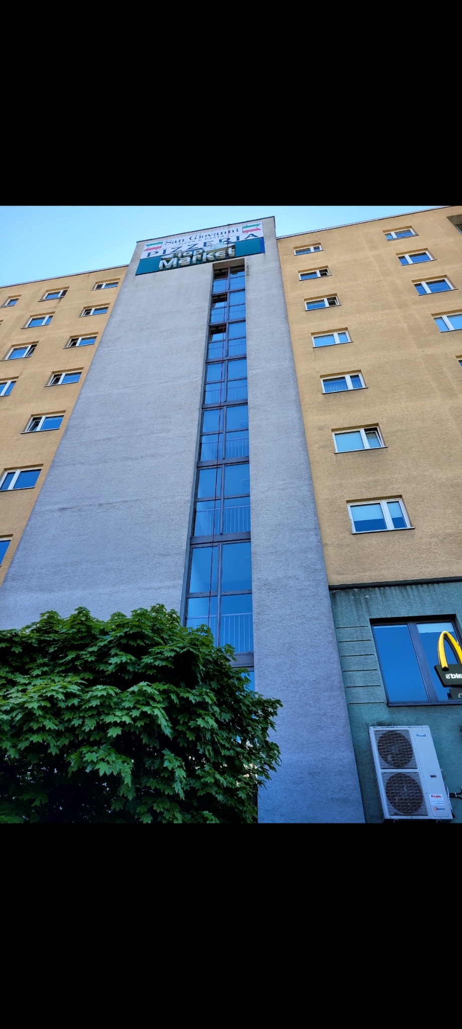 Wysoki budynek z wieloma oknami, z widoczną reklamą na szczycie i klimatyzacją na dole, sfotografowany z dołu.