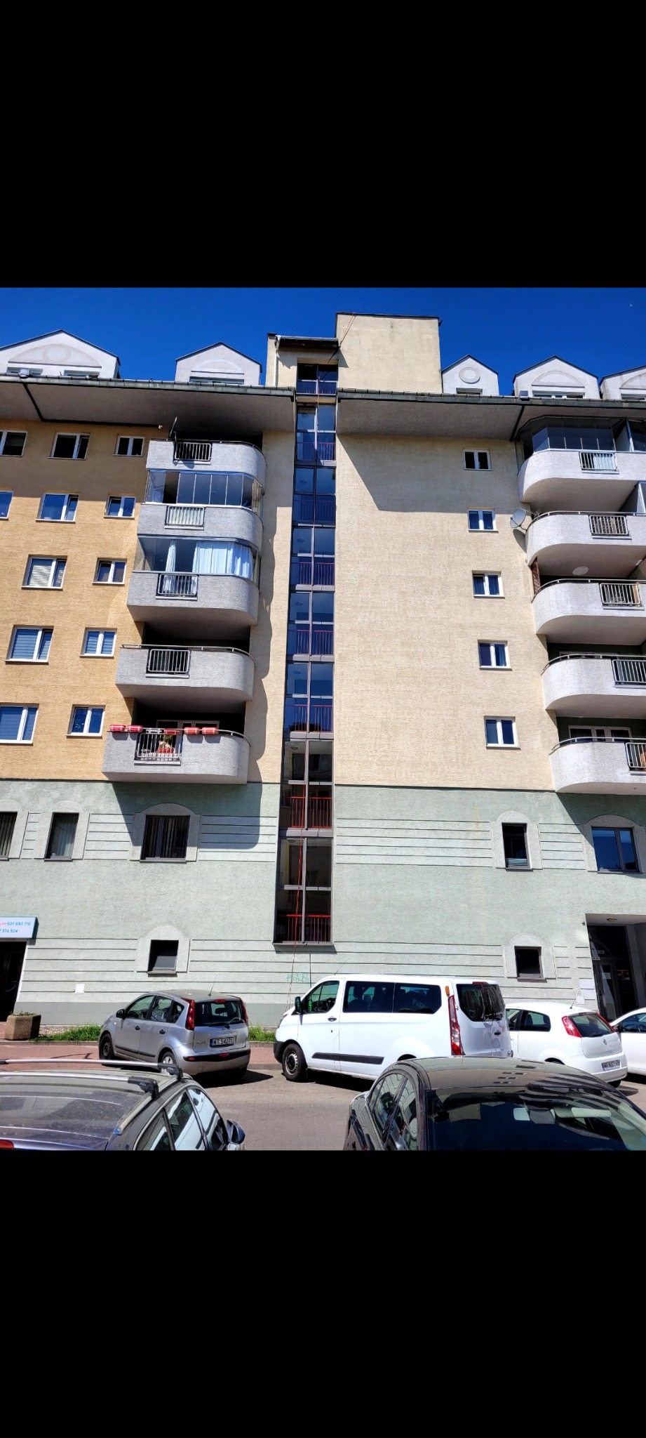 Wielopiętrowy budynek mieszkalny z balkonami i przeszkloną klatką schodową, widoczny z poziomu ulicy z zaparkowanymi samochodami.