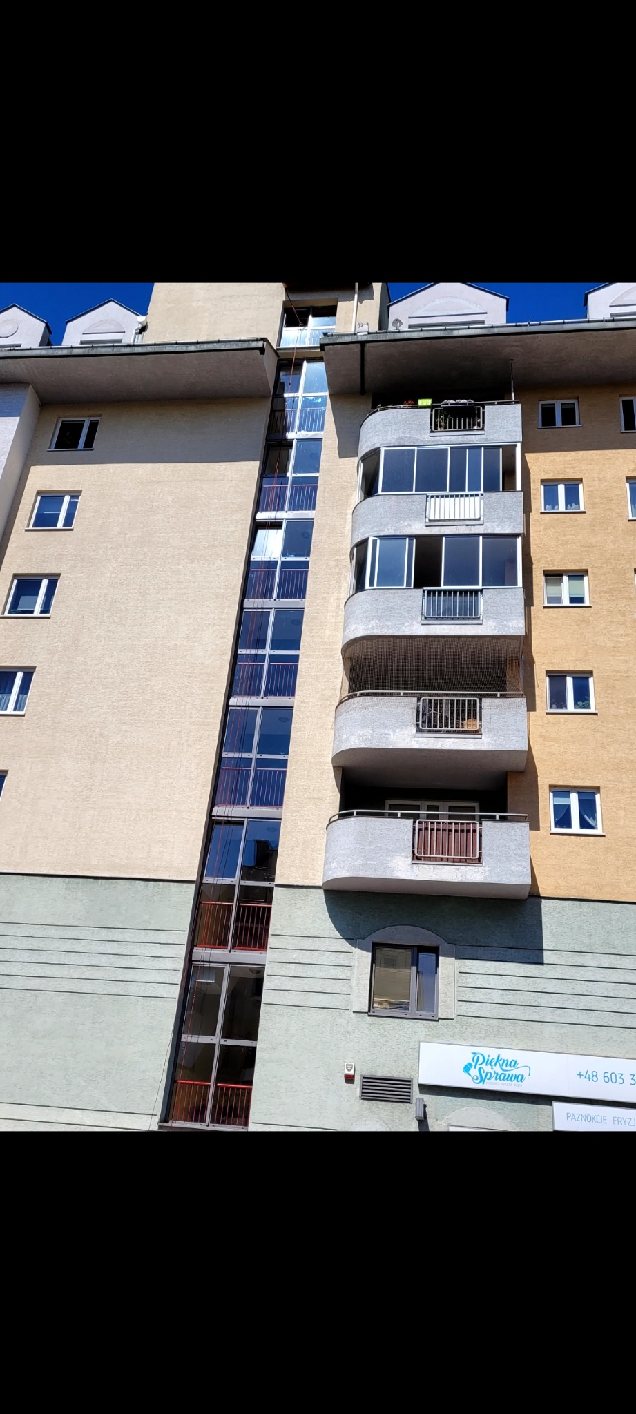 Elewacja budynku wielorodzinnego z przeszkloną klatką schodową, balkonami i szyldem reklamowym salonu kosmetycznego u dołu.