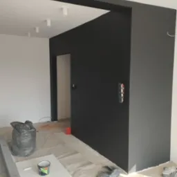 Wnętrze w trakcie malowania na ciemny kolor, zabezpieczona podłoga, widoczne puszki z farbą, wałki i folie malarskie, gniazdko elektryczne bez osłony na ścianie.