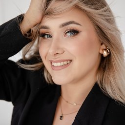 Studio Kreatywne Natalia Kałużyńska - Uśmiechnięta blondynka w czarnej marynarce, z delikatnym makijażem i złotą biżuterią, pozuje z dłonią we włosach na jasnym tle.