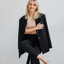 Studio Kreatywne Natalia Kałużyńska - Uśmiechnięta blondynka w czarnym garniturze oversize i beżowych szpilkach siedzi na stołku na tle jasnej ściany, pozując z założonymi rękami.