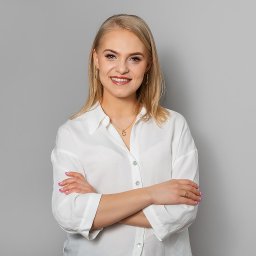 Studio Kreatywne Natalia Kałużyńska - Portret uśmiechniętej blondynki w białej koszuli z podwiniętymi rękawami, skrzyżowanymi rękami, złotym naszyjnikiem z zawieszką misia i złotymi kolczykami na tle szarego studia.