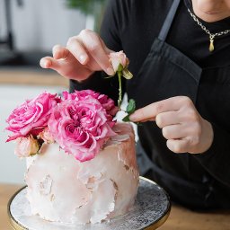 Studio Kreatywne Natalia Kałużyńska - Dekorowanie tortu różami: cukiernik w czarnym fartuchu umieszcza delikatną różową różę na torcie z marmurkową polewą i złotymi akcentami, stojącym na ozdobnym paterze.