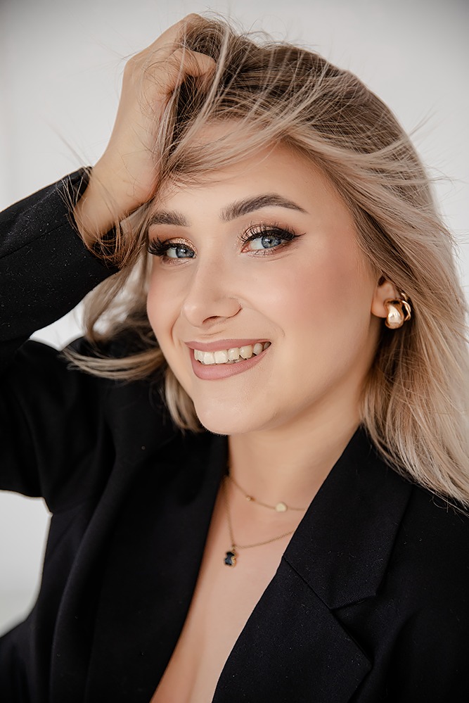 Uśmiechnięta blondynka w czarnej marynarce, z delikatnym makijażem i złotą biżuterią, pozuje z dłonią we włosach na jasnym tle.