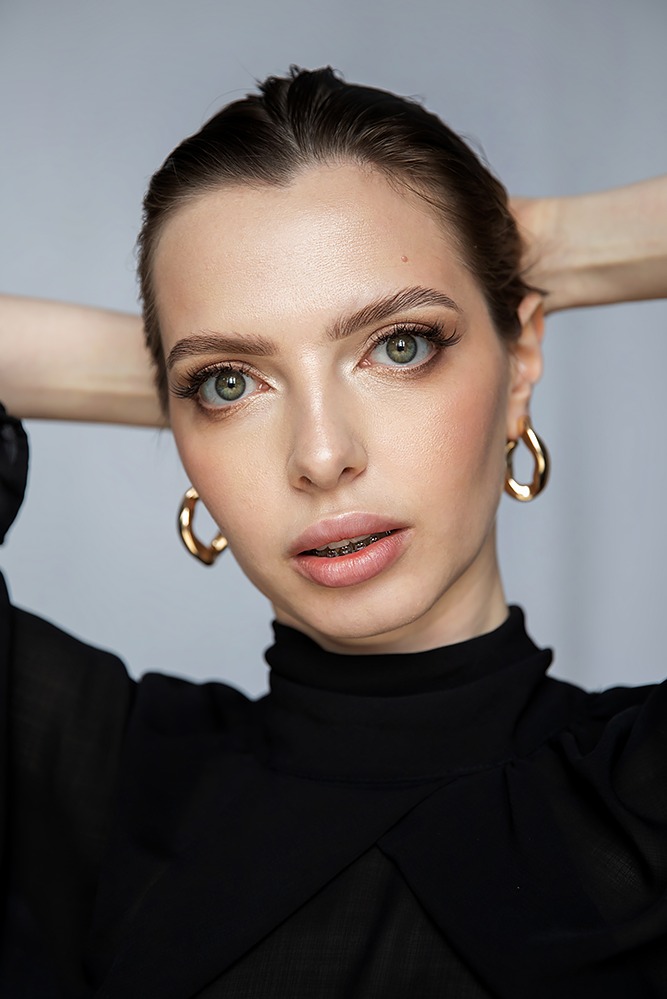 Portret kobiety z brązowymi włosami upiętymi do góry, w czarnej bluzce z golfem, z delikatnym makijażem, złotymi kolczykami i aparatem ortodontycznym na zębach.