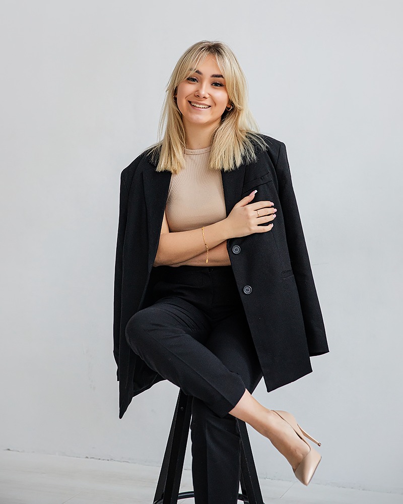 Uśmiechnięta blondynka w czarnym garniturze oversize i beżowych szpilkach siedzi na stołku na tle jasnej ściany, pozując z założonymi rękami.