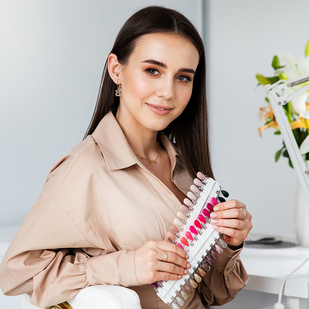 Uśmiechnięta brunetka w beżowej koszuli trzyma paletę lakierów do paznokci z różnymi odcieniami czerwieni i brokatu, widoczna złota biżuteria - kolczyki i naszyjnik.