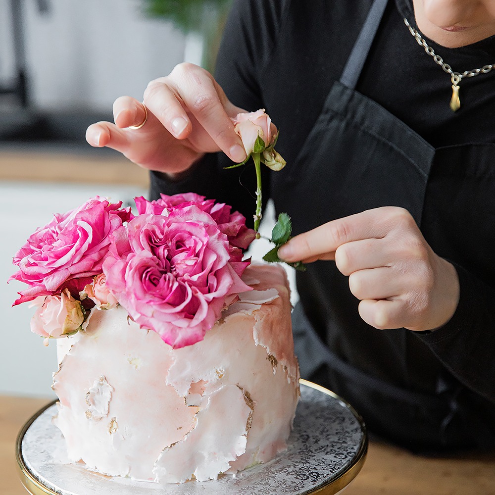 Dekorowanie tortu różami: cukiernik w czarnym fartuchu umieszcza delikatną różową różę na torcie z marmurkową polewą i złotymi akcentami, stojącym na ozdobnym paterze.