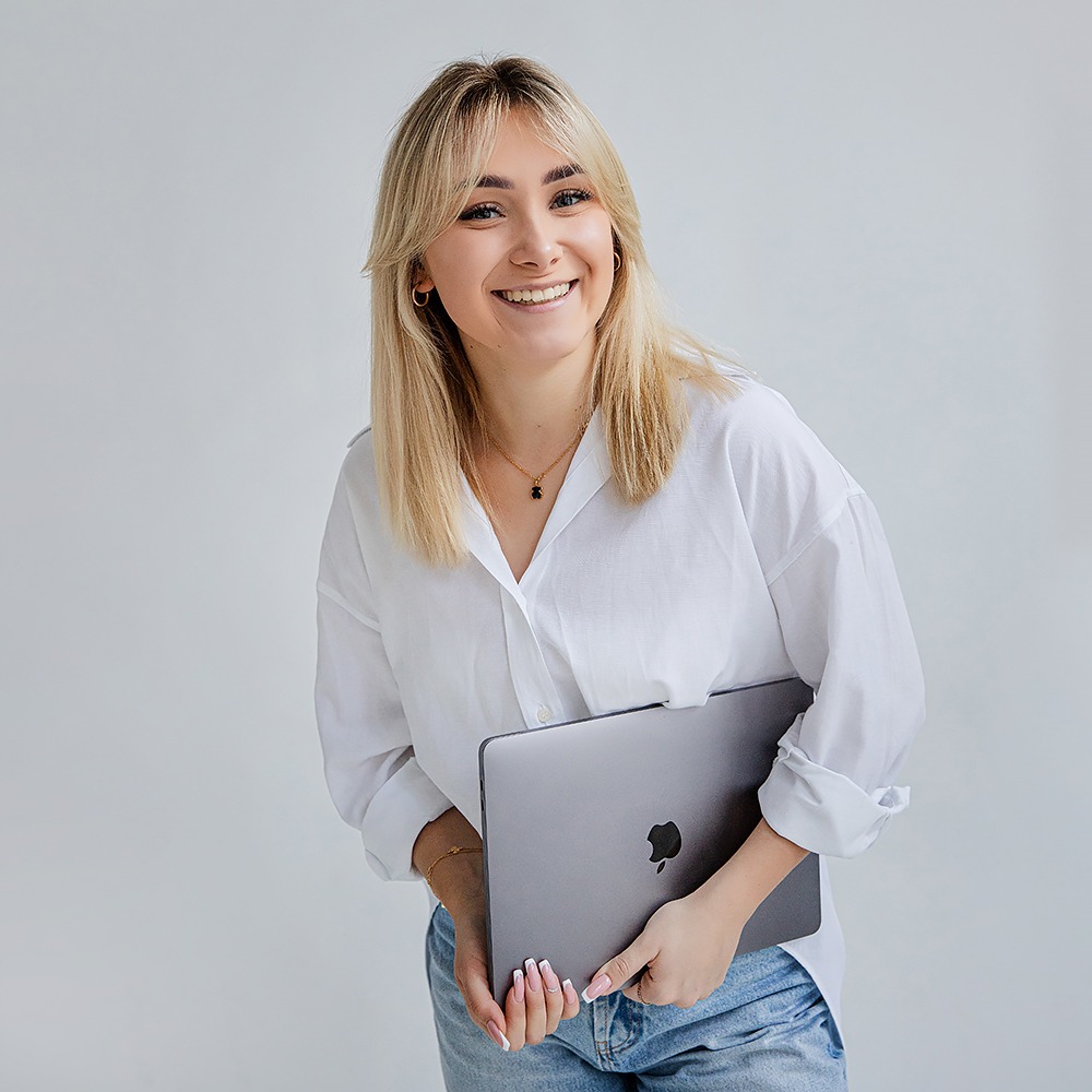 Uśmiechnięta blondynka w białej koszuli i jeansach trzyma zamkniętego laptopa Apple na neutralnym tle.