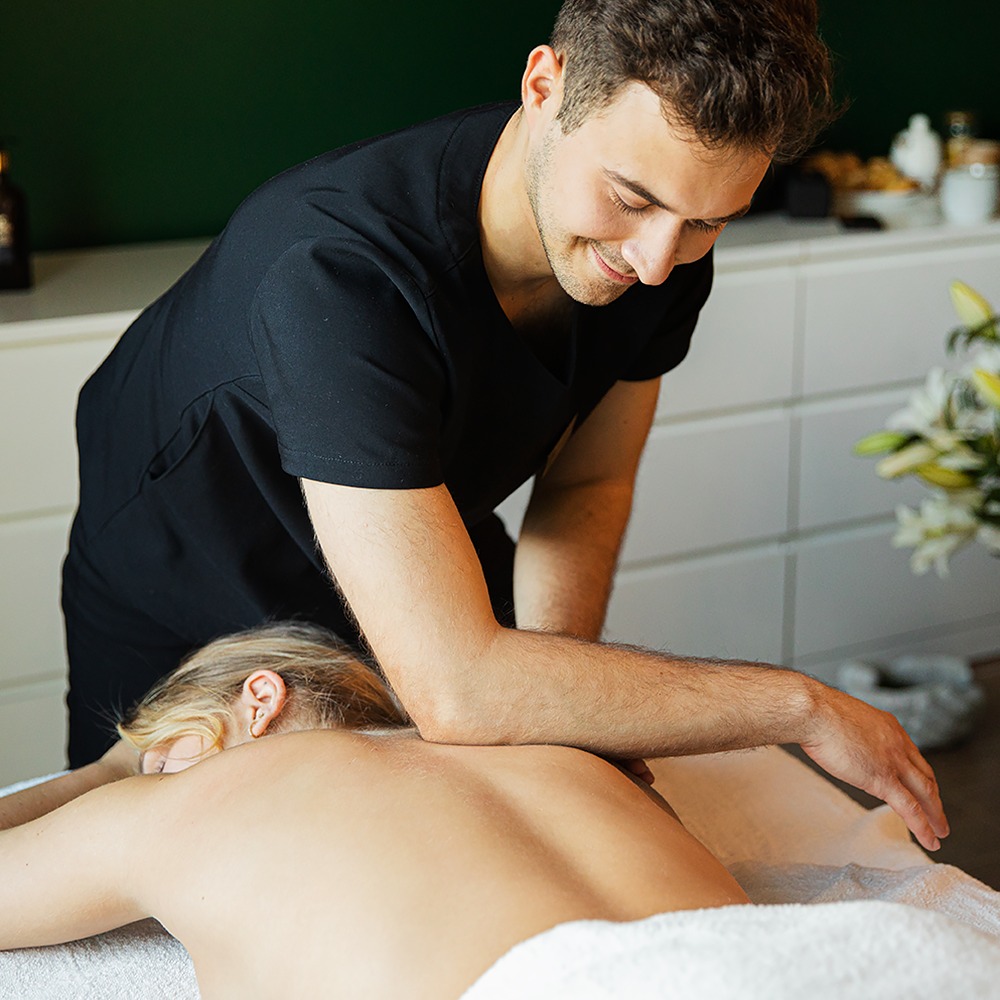 Mężczyzna w czarnym uniformie wykonuje masaż pleców blondynce leżącej na białym ręczniku, w tle biała komoda i bukiet białych lilii.