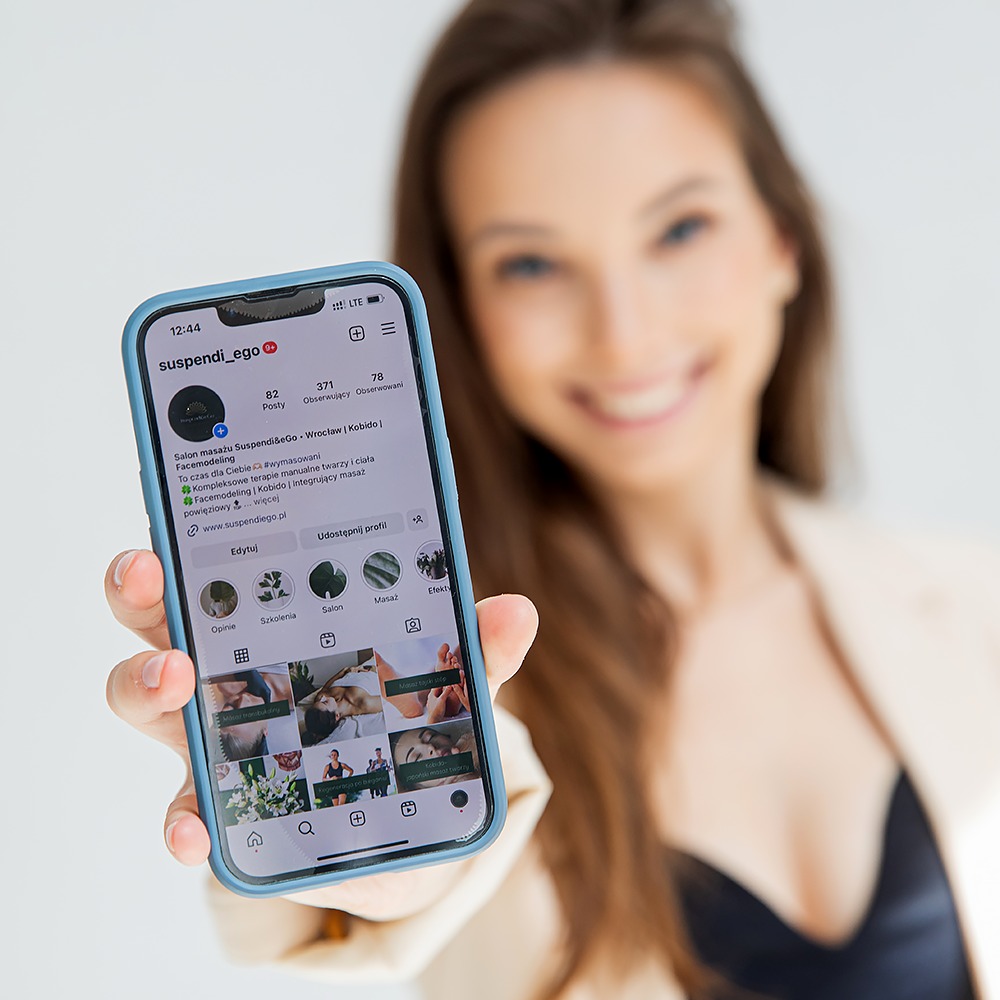 Uśmiechnięta kobieta w beżowej marynarce prezentuje ekran smartfona z profilem salonu masażu Suspendi&eGo, oferującego zabiegi Kobido i Facemodeling.