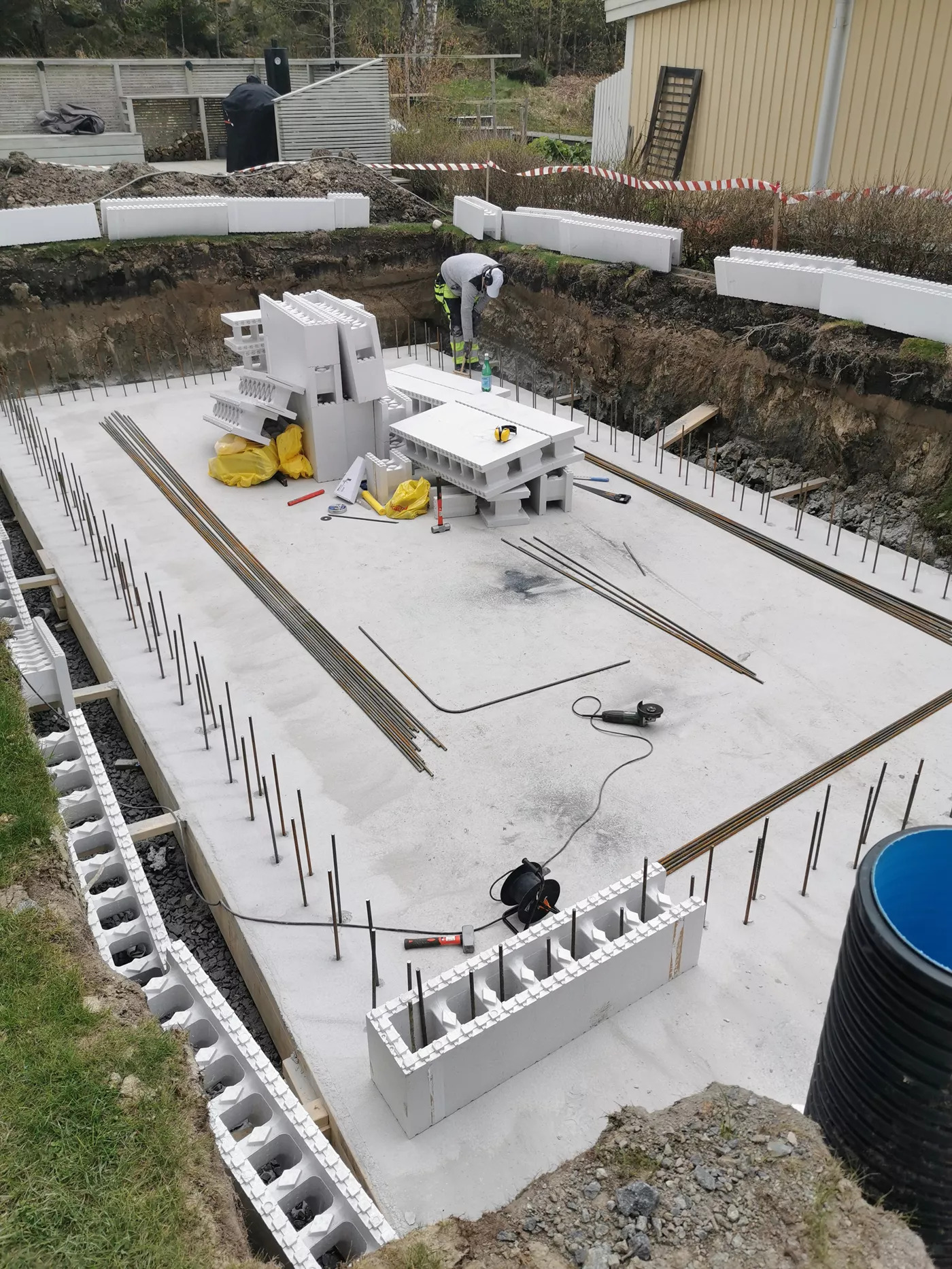 Przygotowanie fundamentów pod budowę, widoczne szalunki z białych bloczków styropianowych, wystające pręty zbrojeniowe, narzędzia budowlane i pracownik w tle.