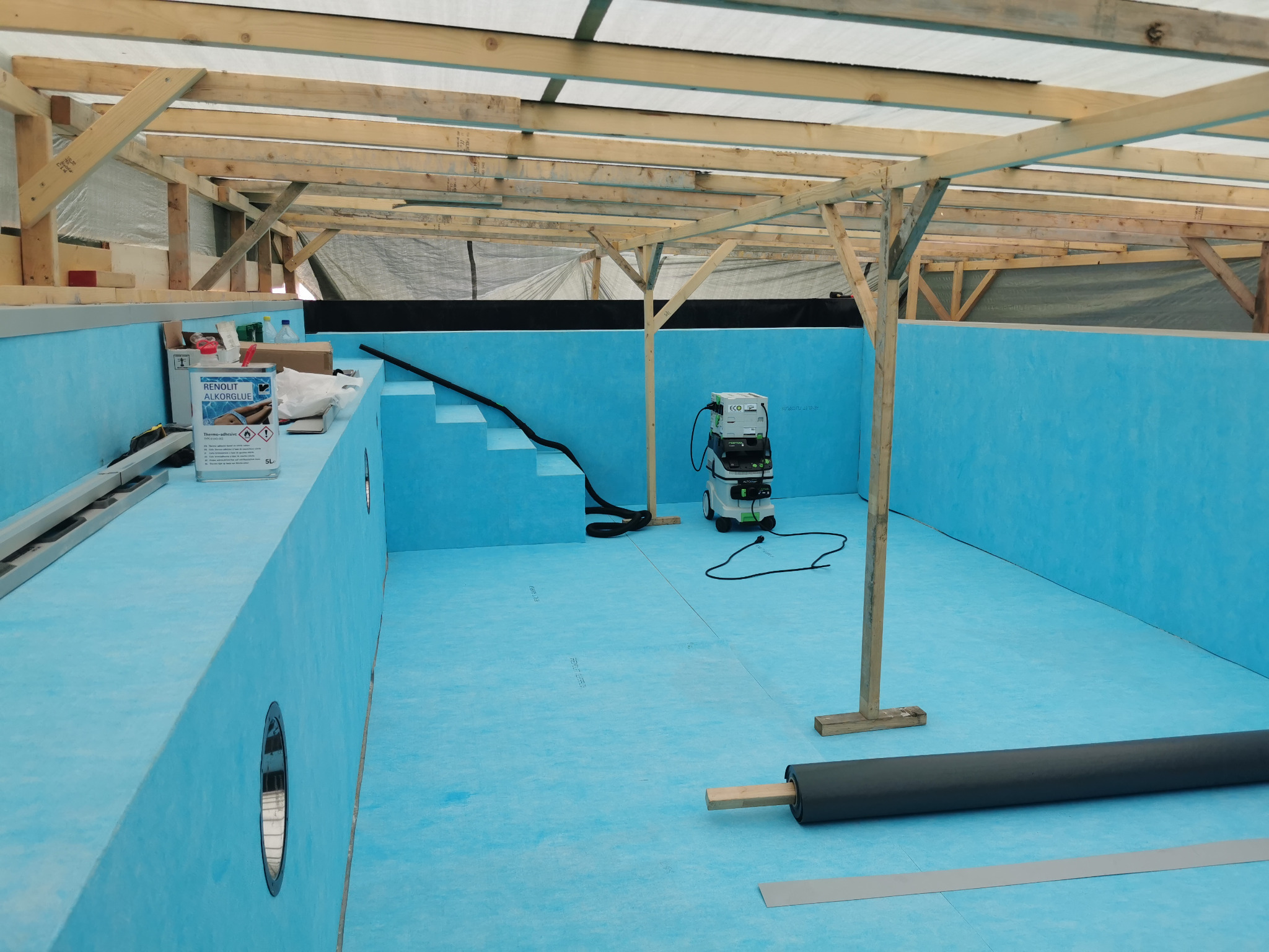 Wnętrze budowanego basenu z niebieską folią, widoczny szkielet konstrukcji z drewna, rolka folii, odkurzacz przemysłowy i puszka kleju.