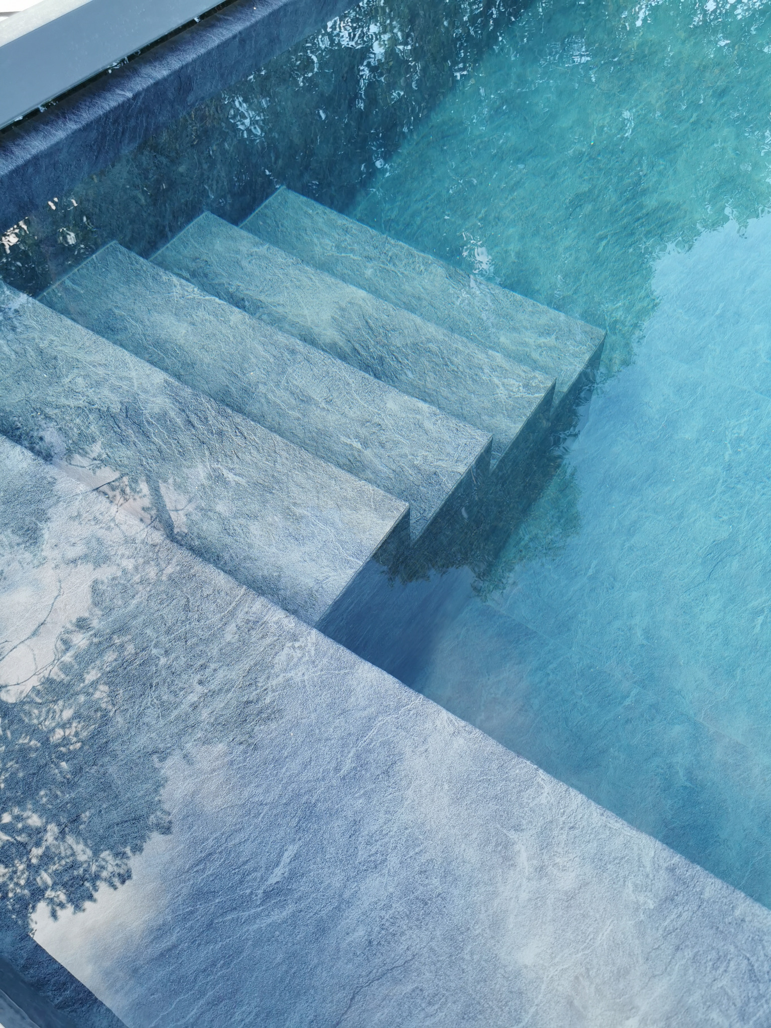 Schody z szarego kamienia prowadzące do krystalicznie czystej, błękitnej wody w basenie, z refleksami drzew na powierzchni.