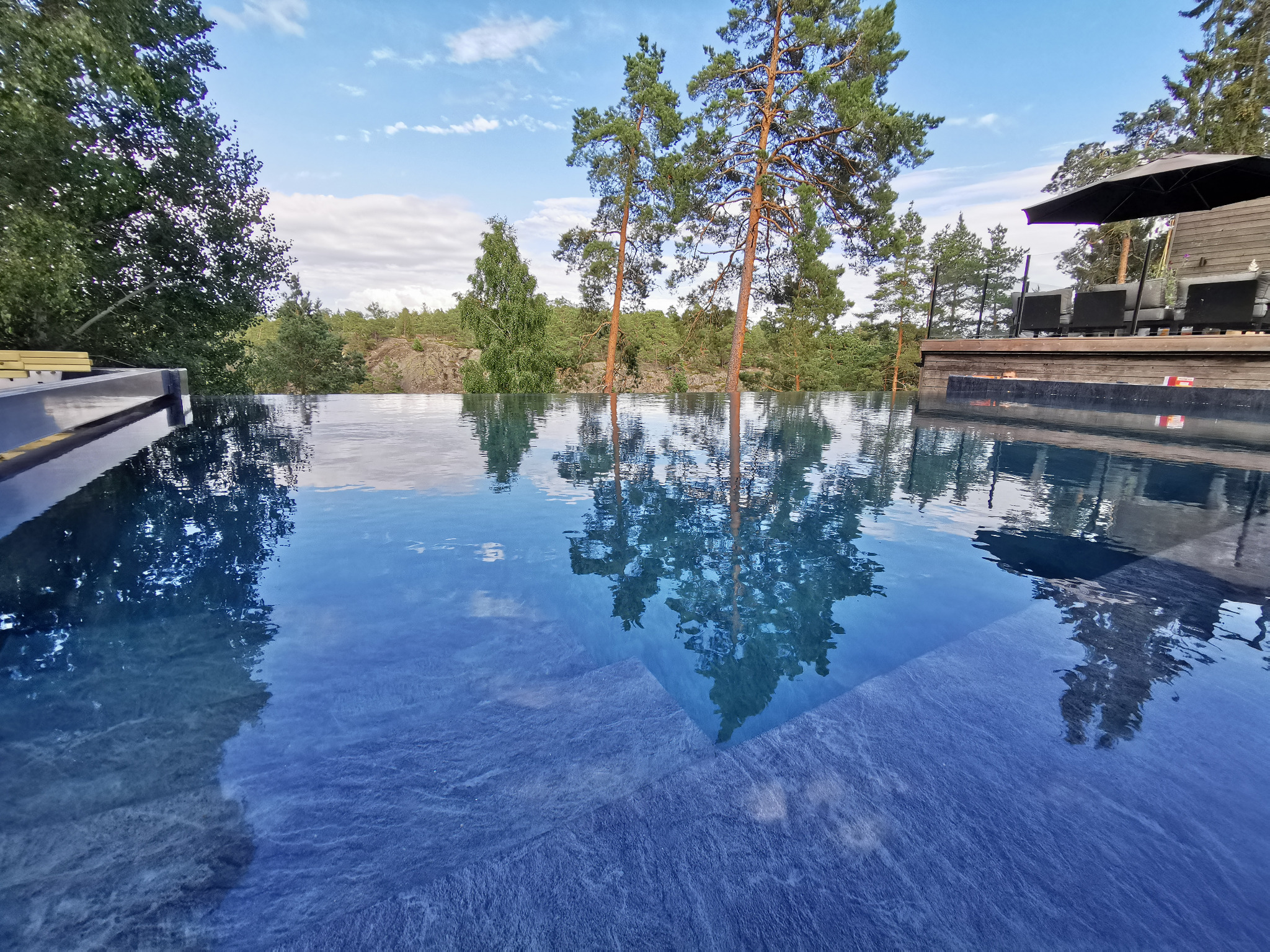 Nowoczesny basen bez krawędzi z ciemnoniebieską wodą i kamiennym wykończeniem, odbijający drzewa i błękitne niebo, widok z poziomu wody.