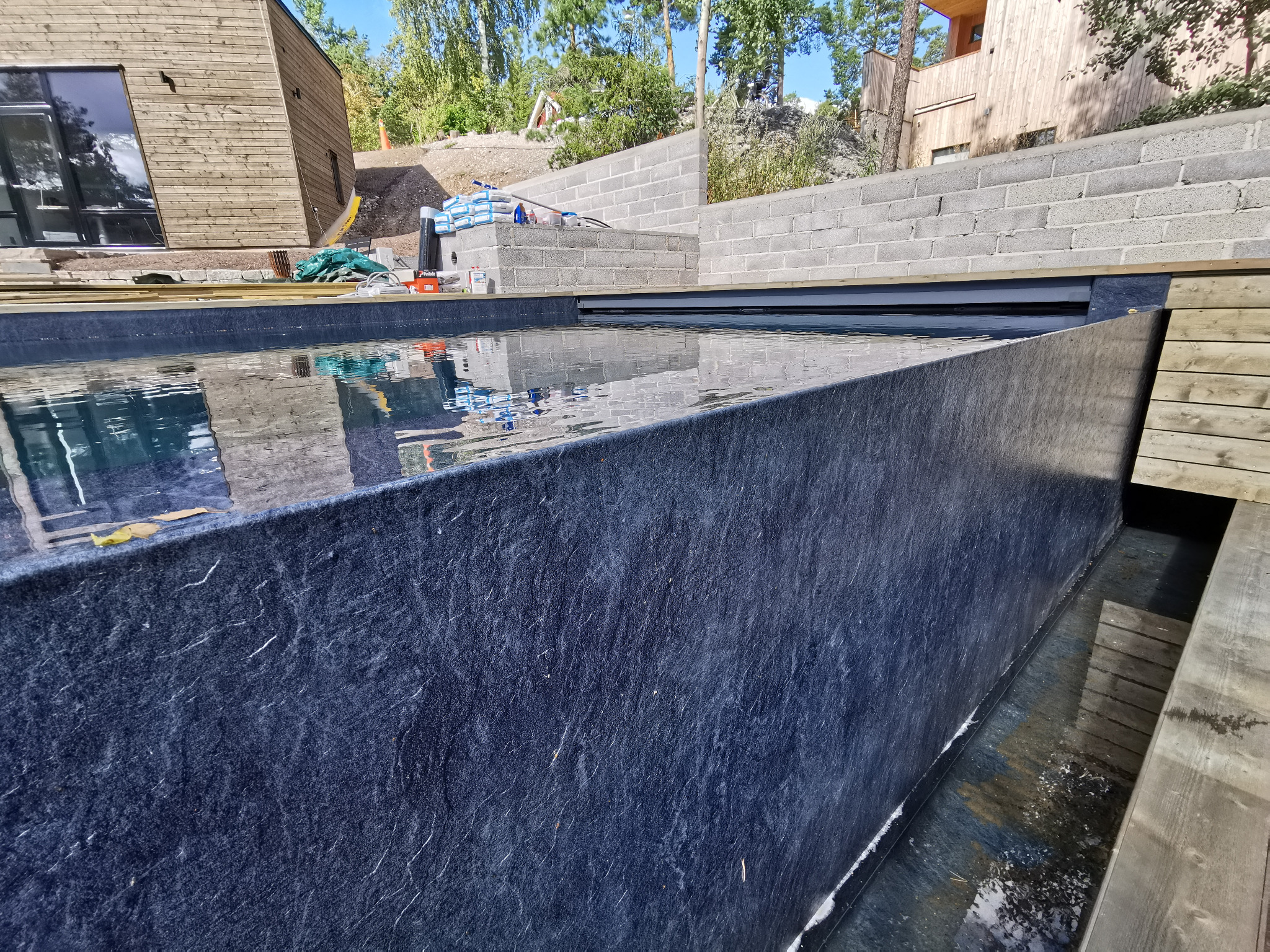 Nowoczesny basen w trakcie budowy, wykończony ciemnym kamieniem, z widocznymi bloczkami betonowymi w tle i elementami drewnianymi.