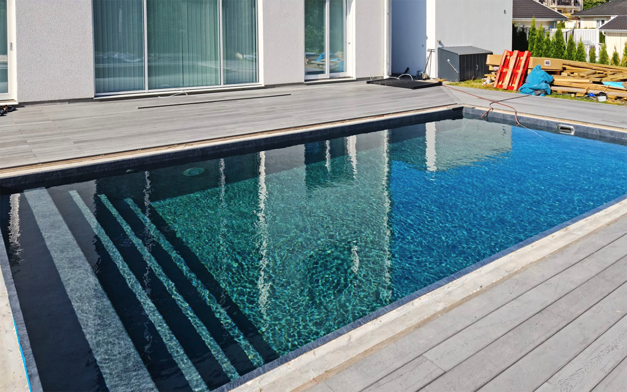 Nowoczesny basen z szarymi schodami i drewnianym tarasem, w tle widoczne elementy budowy domu i materiały budowlane.