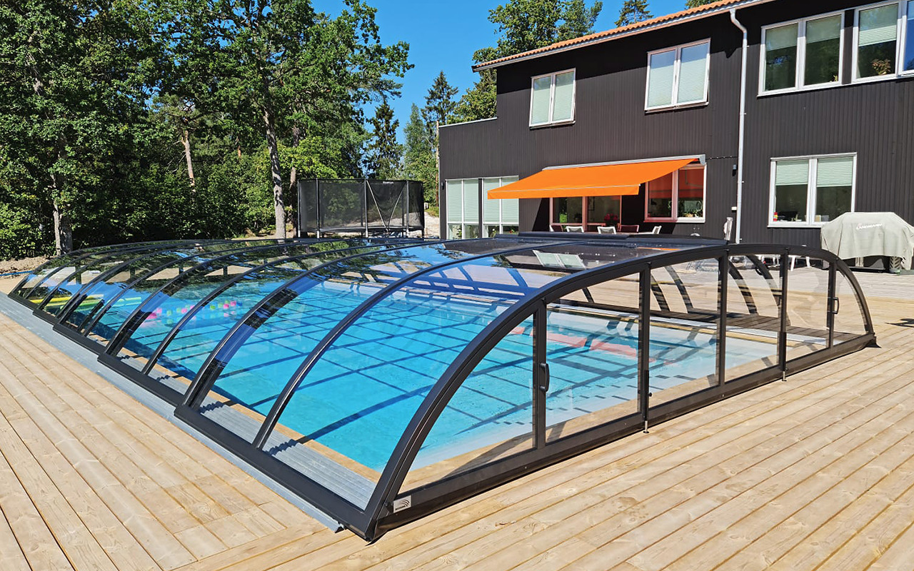 Zadaszony basen ogrodowy z przesuwnymi segmentami, otoczony drewnianym tarasem, na tle ciemnego domu z pomarańczową markizą i zieleni drzew.