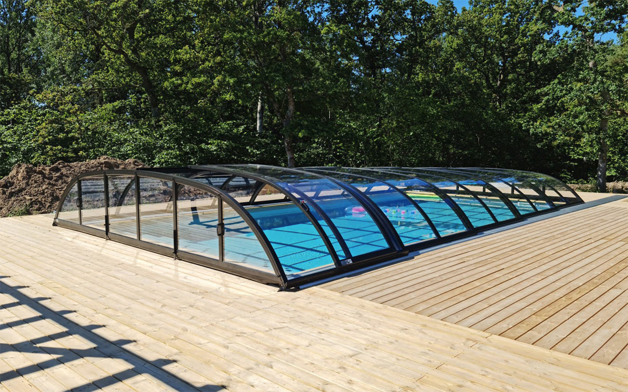 Zadaszony basen zewnętrzny z czarną ramą i przezroczystymi panelami, otoczony drewnianym tarasem, z widocznym stosem ziemi w tle i zielenią drzew.