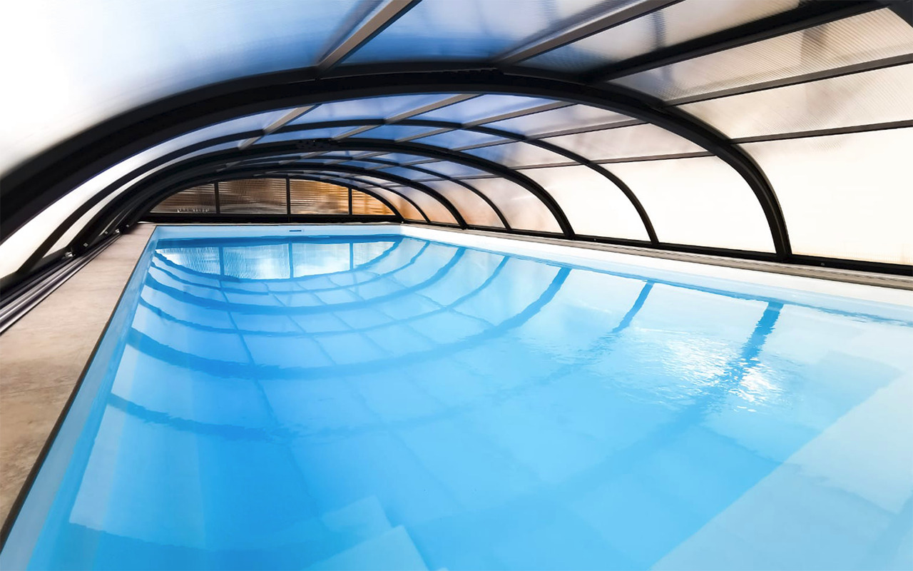 Kryty basen z przezroczystym zadaszeniem łukowym z czarną ramą, odbicie nieba w tafli wody.