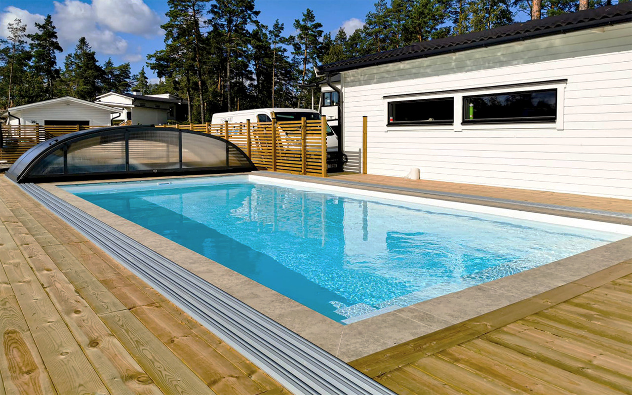 Prostokątny, zewnętrzny zbiornik wodny z przejrzystą, przesuwną osłoną, otoczony drewnianym tarasem, z widocznymi domami i drzewami w tle.