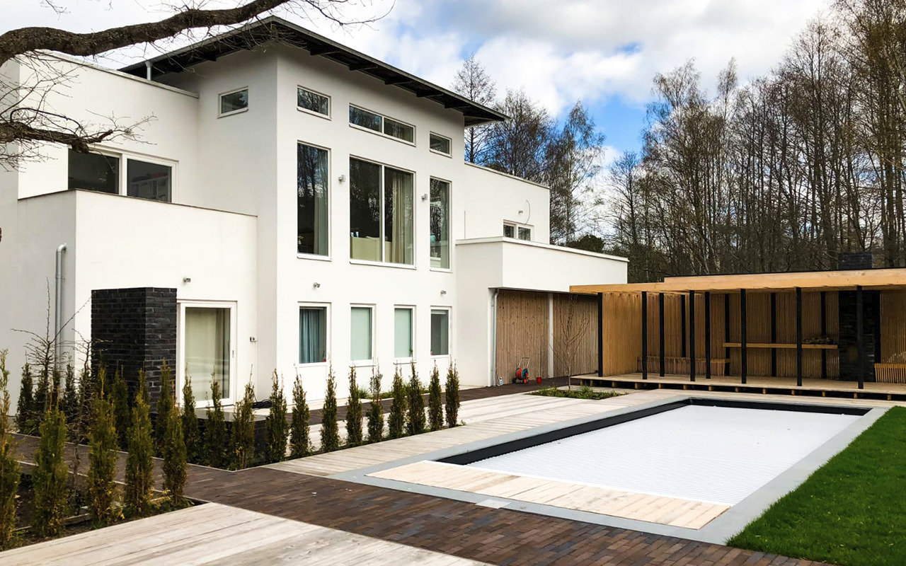 Nowoczesny dom z białymi ścianami, zadaszonym tarasem z drewna i prostokątnym basenem z roletą, otoczony kostką brukową i pasem zieleni.