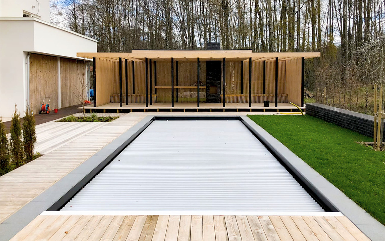 Zadaszony basen z roletą zabezpieczającą, obok drewniana altana z grillem i trawnik