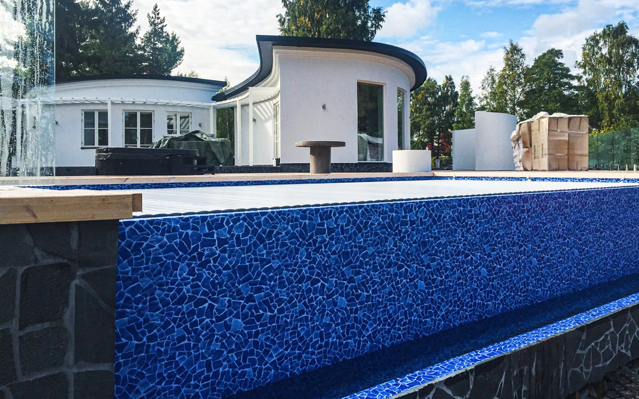 Niebieska mozaika wykończeniowa na ścianach basenu, w tle nowoczesny dom z zaokrąglonymi elementami i częściowo zakryta wanna SPA.