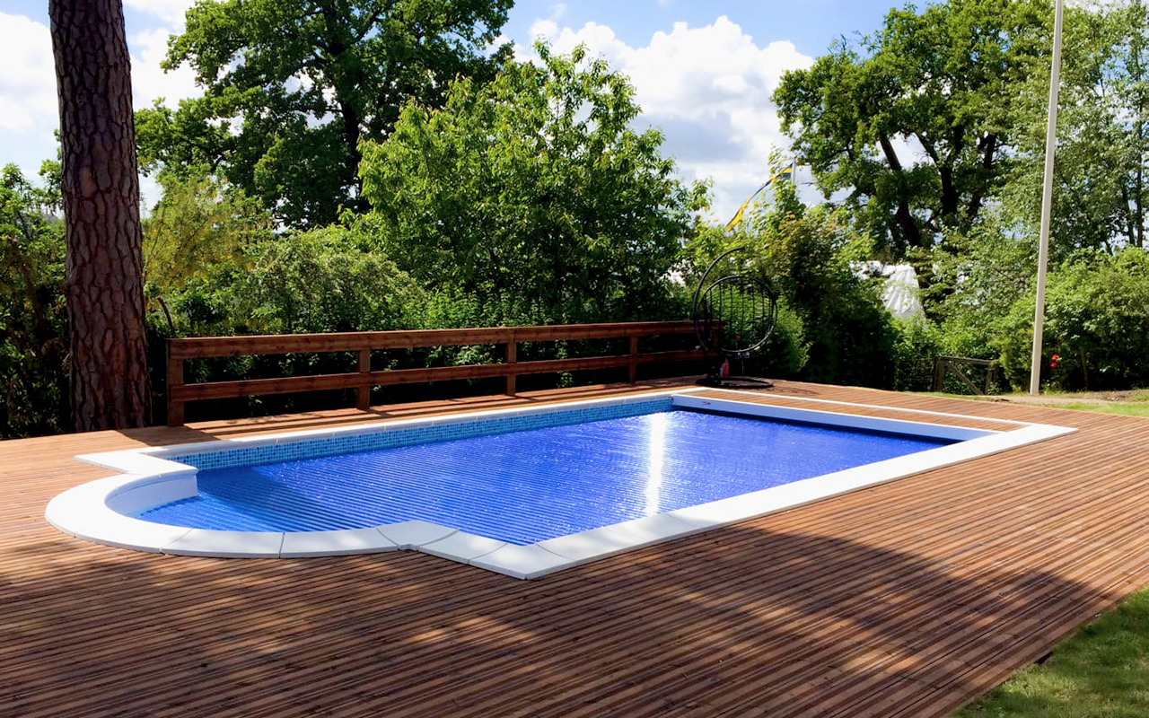 Kryty basen ogrodowy z drewnianym tarasem i widocznym drzewem obok, otoczony zielenią.