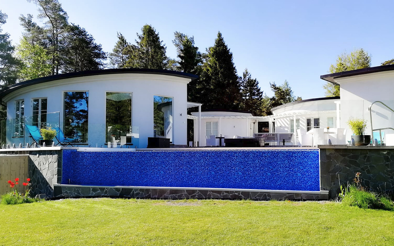 Nowoczesny, częściowo ukryty basen z mozaiką w kolorze niebieskim, otoczony zielonym trawnikiem i białymi, okrągłymi budynkami z dużymi oknami.