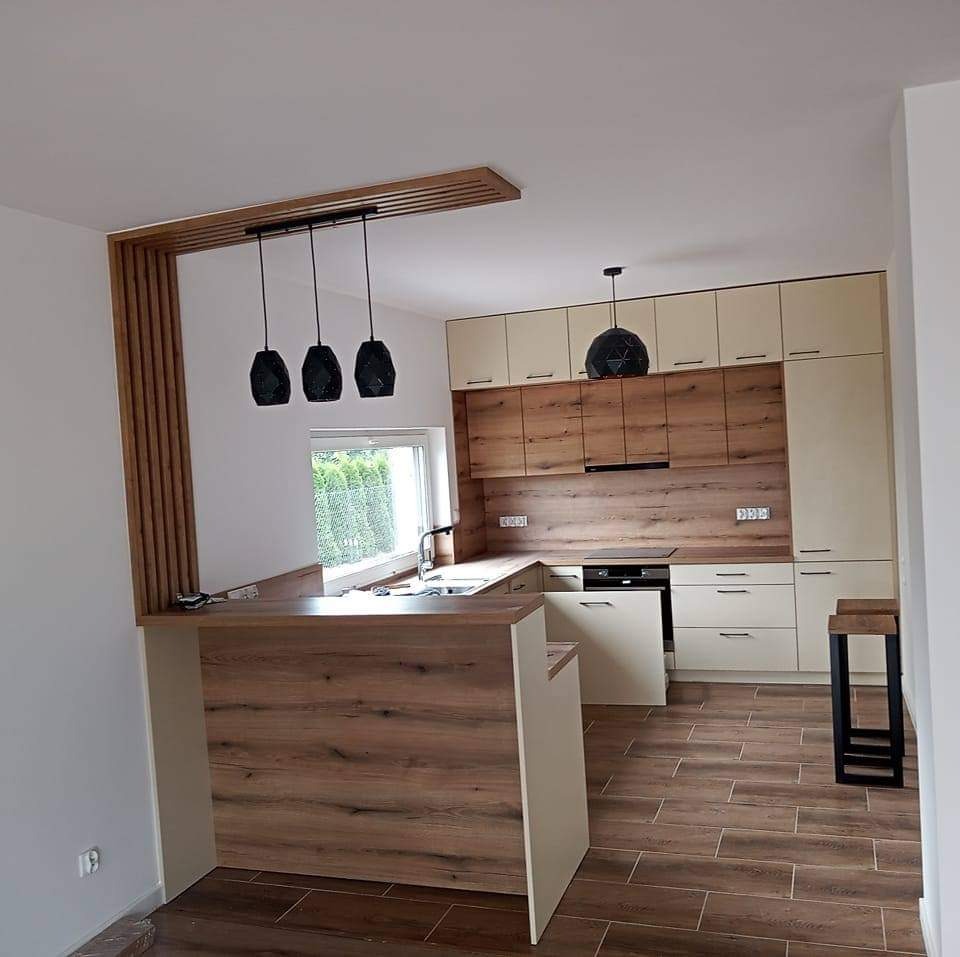 Nowoczesna kuchnia z drewnianymi akcentami: blat baru, szafki dolne i górne w odcieniach kremu i drewna, trzy czarne lampy wiszące nad barem, podłoga z płytek imitujących drewno, okno z widokiem...