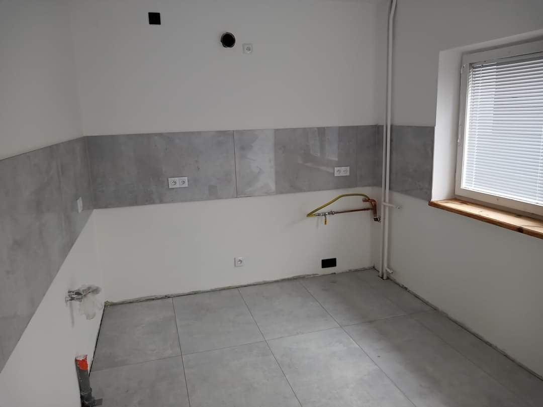 Surowe wnętrze kuchni w trakcie remontu: szare płytki na podłodze i ścianie, odkryte instalacje wodne, gładkie białe ściany, okno z żaluzjami.