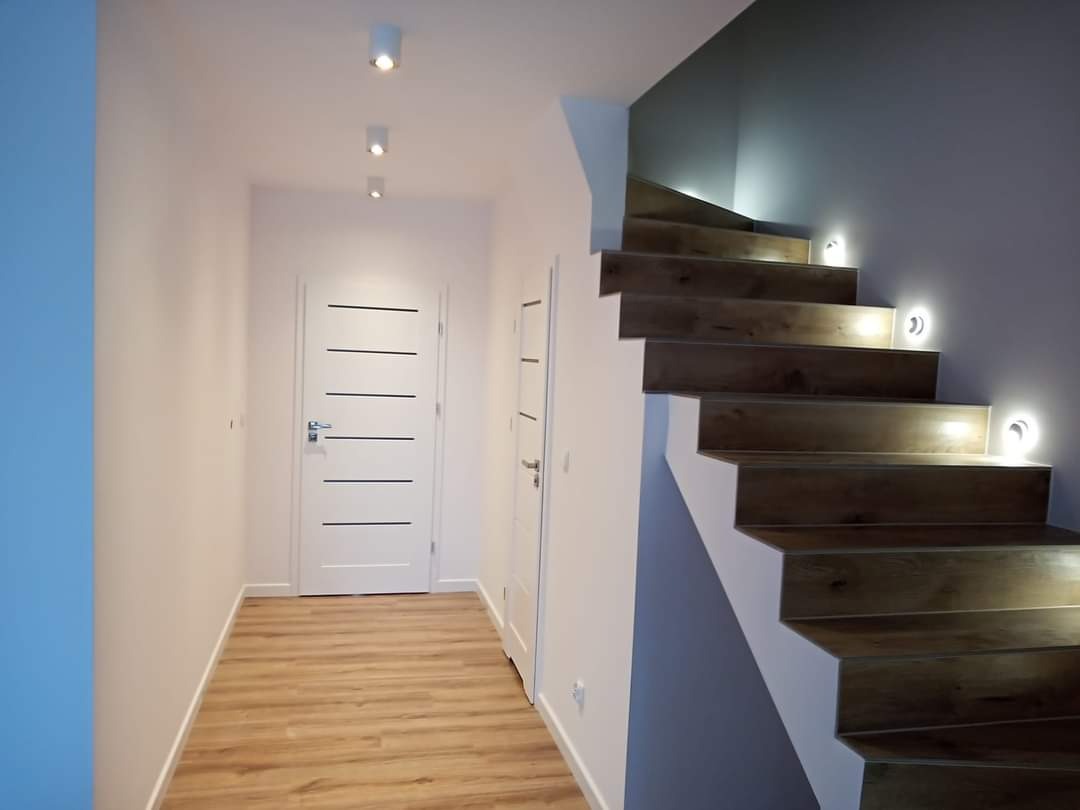 Nowoczesny przedpokój z drewnopodobnymi panelami na podłodze i schodach, oświetlenie punktowe sufitowe i schodowe, białe drzwi z poziomymi, czarnymi paskami.
