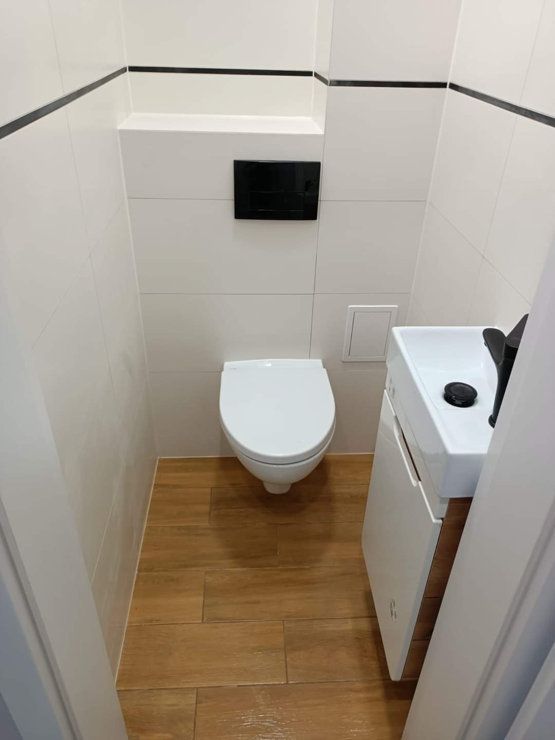 Wąska toaleta z białymi płytkami, drewnopodobną podłogą, wiszącą muszlą klozetową i małą umywalką z szafką, wykończona czarnymi akcentami na ścianach.