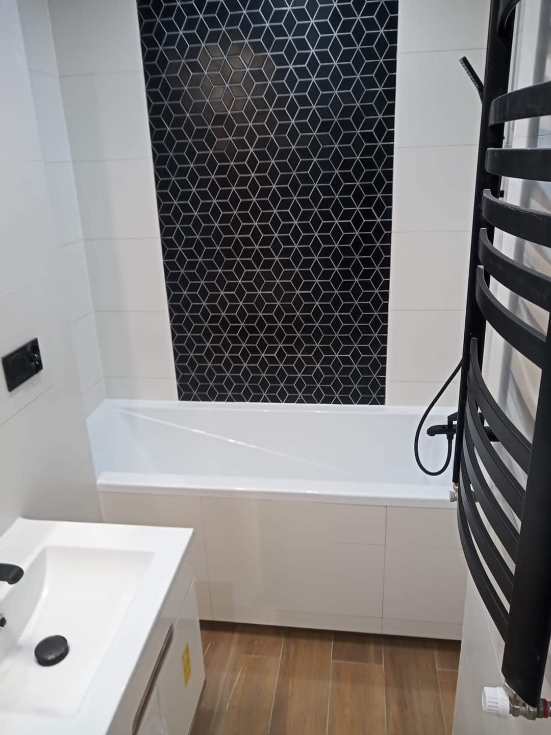 Nowoczesna łazienka z białą wanną obudowaną panelami, umywalką z szafką i czarnym grzejnikiem drabinkowym; na ścianie białe płytki oraz czarny pas z geometrycznym wzorem.