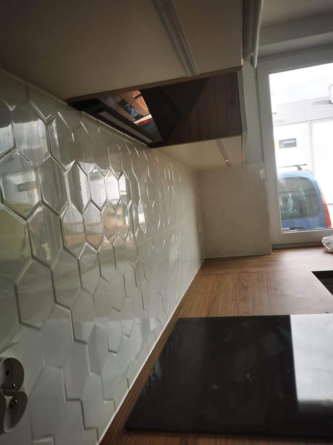 Białe, heksagonalne płytki na ścianie w kuchni, z widocznym fragmentem blatu i płyty indukcyjnej oraz szafki wiszącej z oświetleniem.