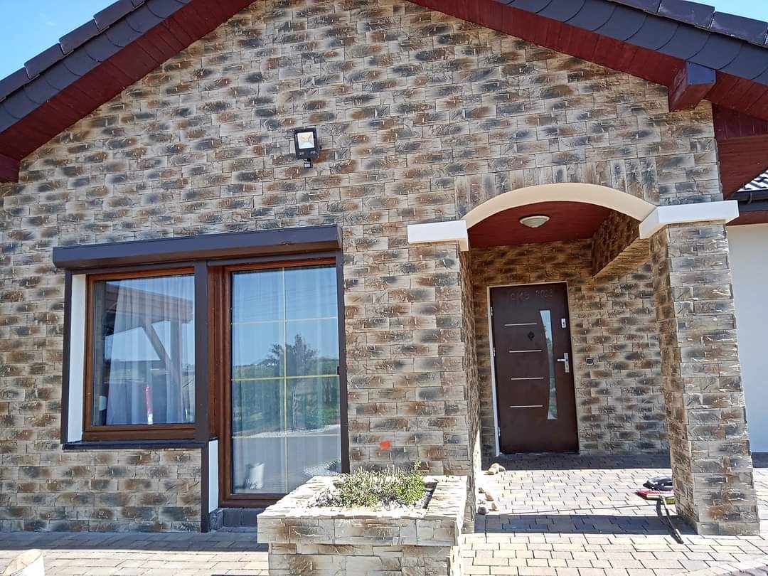 Elewacja domu jednorodzinnego wykończona dekoracyjną cegłą w odcieniach szarości i brązu, widoczne okno z brązową ramą i roletą zewnętrzną, drzwi wejściowe z zadaszeniem i kolumnami, oraz fragment...