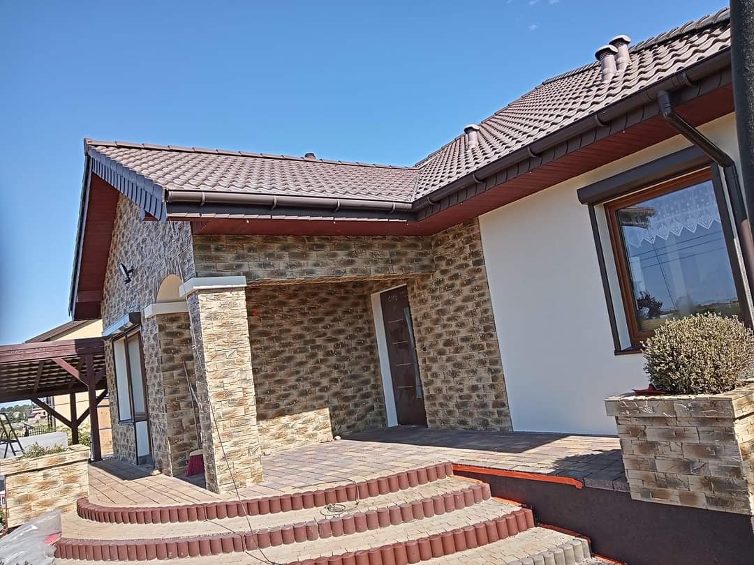 Elewacja domu z kamienia dekoracyjnego, brązowy dach z dachówki ceramicznej, okno z roletą zewnętrzną, schody wejściowe wyłożone kostką brukową w odcieniach brązu i beżu.