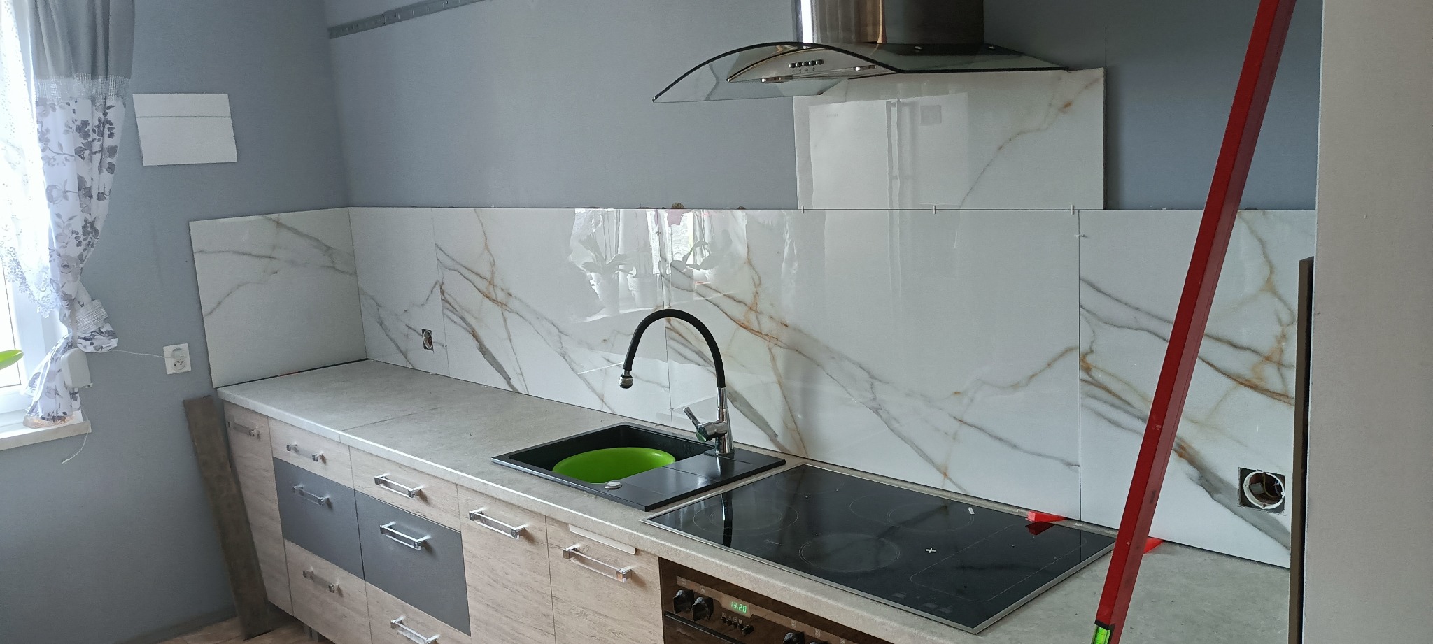 Montaż płytek imitujących marmur na ścianie w kuchni, widoczny fragment blatu, zlew z zieloną miską, płyta indukcyjna i poziomica oparta o ścianę.