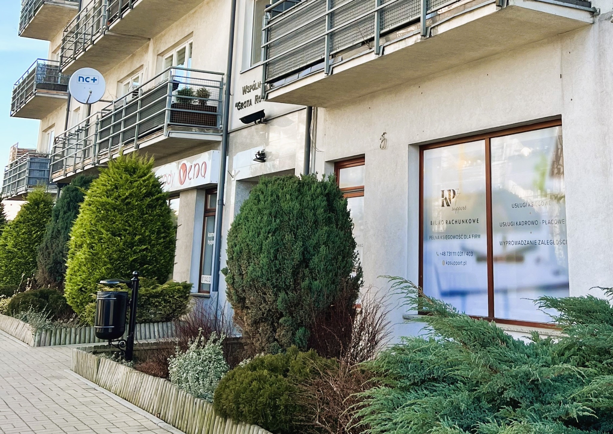 Fasada budynku w Pabianicach z widocznym oknem biura rachunkowego KP Support oferującego usługi księgowe i kadrowe, przysłonięta częściowo przez krzewy.